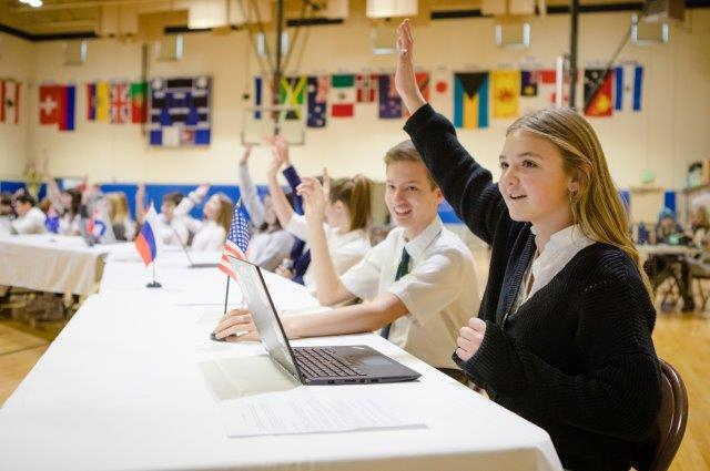 Students at a table with computers raising their hands. Country flags on the walls in the background..